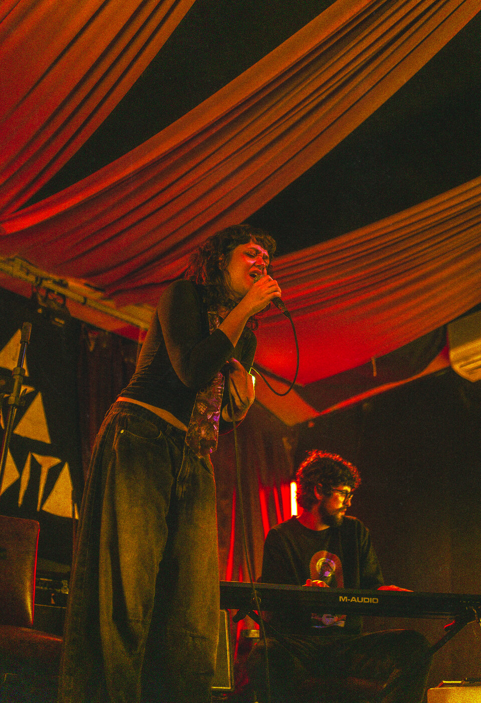 BRUNÊ cantando ao microfone num palco pequeno, cortina vermelha ao fundo