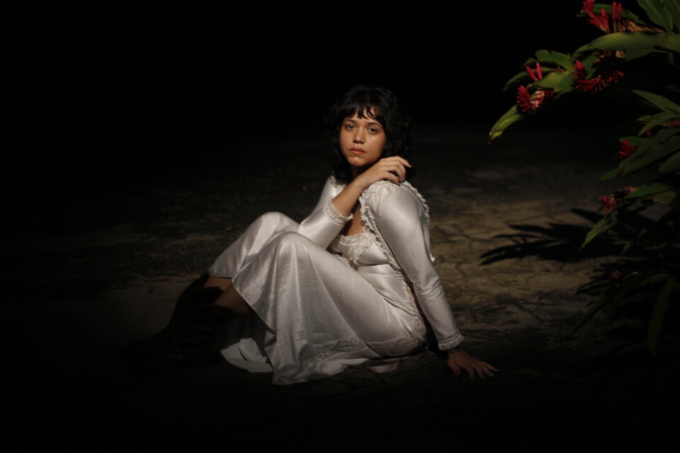 BRUNÊ sentada no chão à noite, de vestido claro, ao lado de uma flor tropical